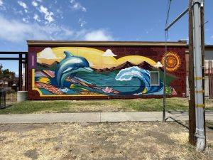 A colorful outdoor mural painted on a school building wall. It shows a blue dolphin on the left, a breaking wave on the right, a pink paper boat between them, and hills under a sun in the background. Caption: "Two Languages, Un Corazón" by Richard Muro Salazar. ARTSCCC photo.