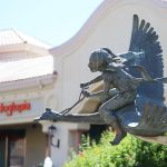 Gallery 1 - A bronze sculpture shows a young girl with pigtails holding reins as she rides on the back of a flying bird. The piece is set outdoors against a backdrop of trees and a shopping plaza.