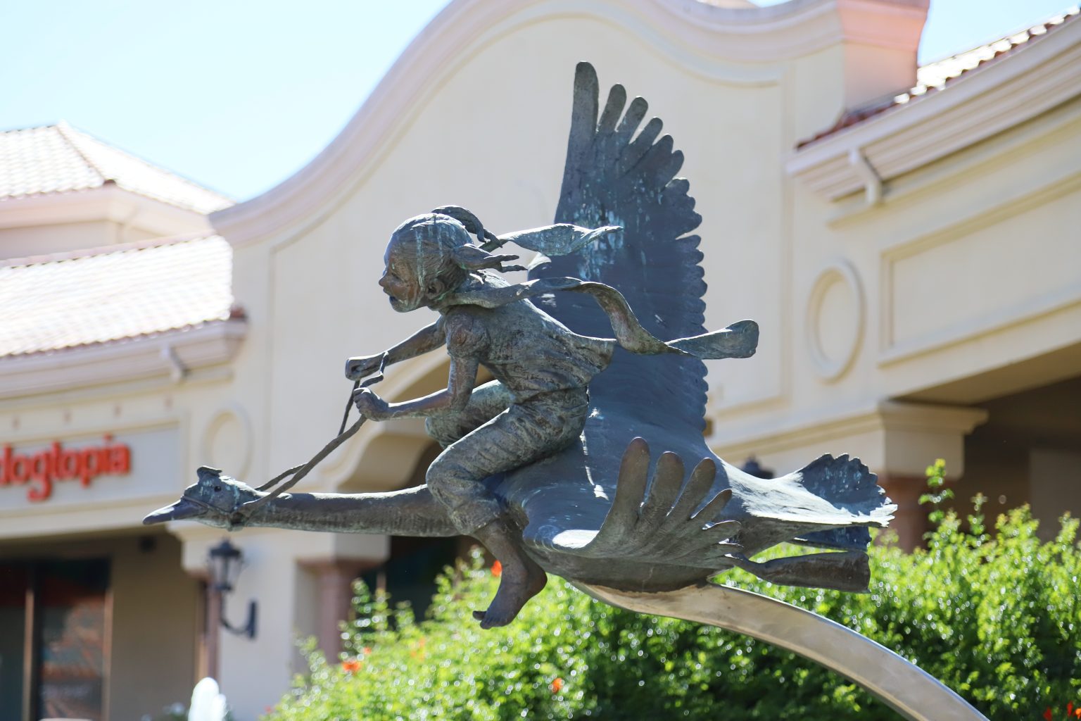 Gallery 1 - A bronze sculpture shows a young girl with pigtails holding reins as she rides on the back of a flying bird. The piece is set outdoors against a backdrop of trees and a shopping plaza.
