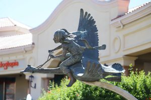 A bronze sculpture shows a young girl with pigtails holding reins as she rides on the back of a flying bird. The piece is set outdoors against a backdrop of trees and a shopping plaza.