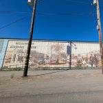 Gallery 1 - A wide outdoor mural painted in sepia tones across corrugated metal siding shows historical buildings, a palm tree, and a vintage steam train with arriving passengers, alongside text reading 