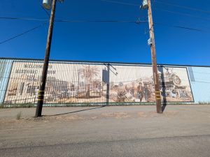 A wide outdoor mural painted in sepia tones across corrugated metal siding shows historical buildings, a palm tree, and a vintage steam train with arriving passengers, alongside text reading "Welcome to Historic Byron Est. 1878 by Tom Preis."