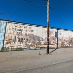 Gallery 2 - A wide outdoor mural painted in sepia tones across corrugated metal siding shows historical buildings, a palm tree, and a vintage steam train with arriving passengers, alongside text reading 