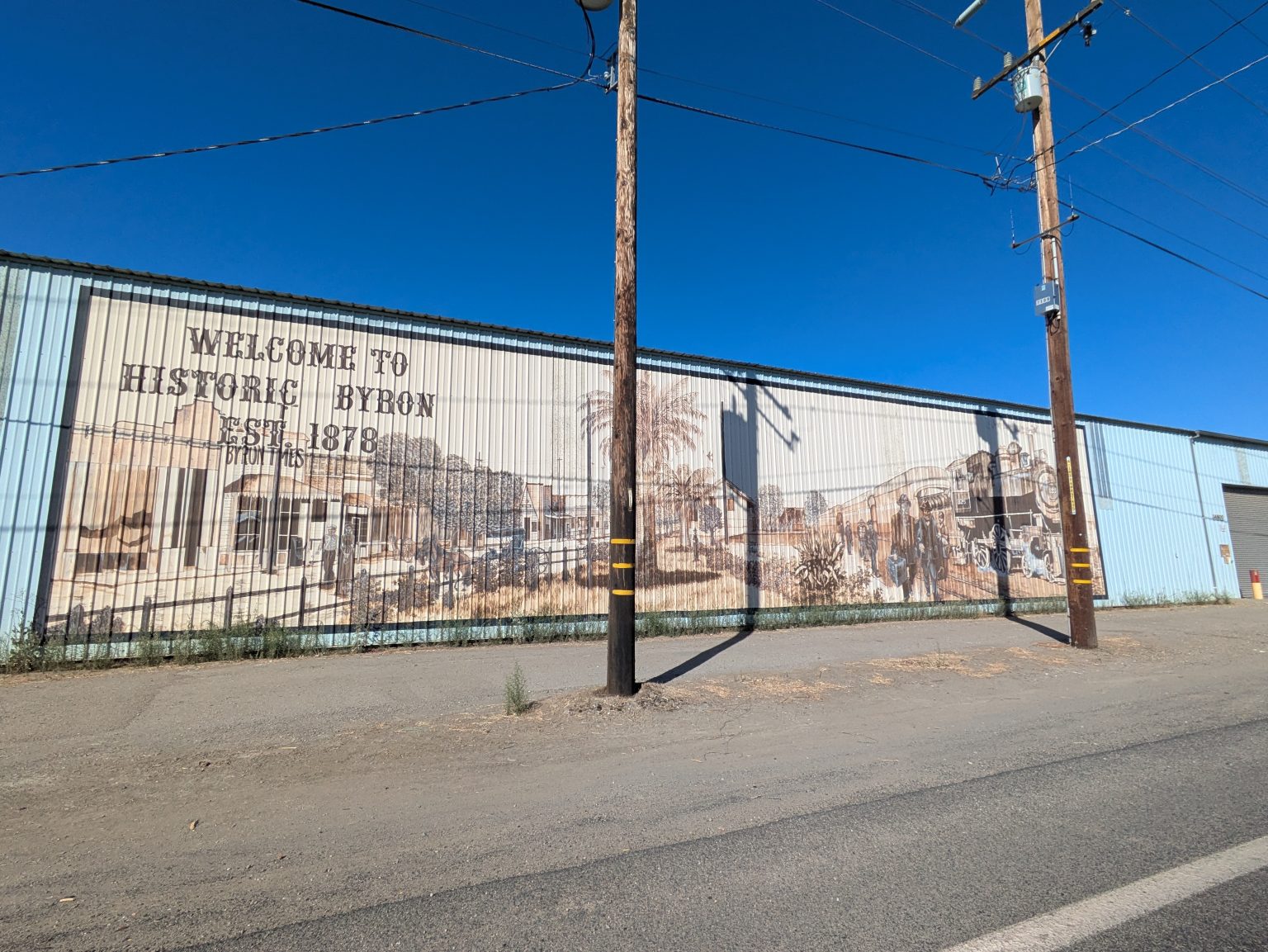 Gallery 2 - A wide outdoor mural painted in sepia tones across corrugated metal siding shows historical buildings, a palm tree, and a vintage steam train with arriving passengers, alongside text reading 