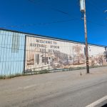 Gallery 3 - A wide outdoor mural painted in sepia tones across corrugated metal siding shows historical buildings, a palm tree, and a vintage steam train with arriving passengers, alongside text reading 