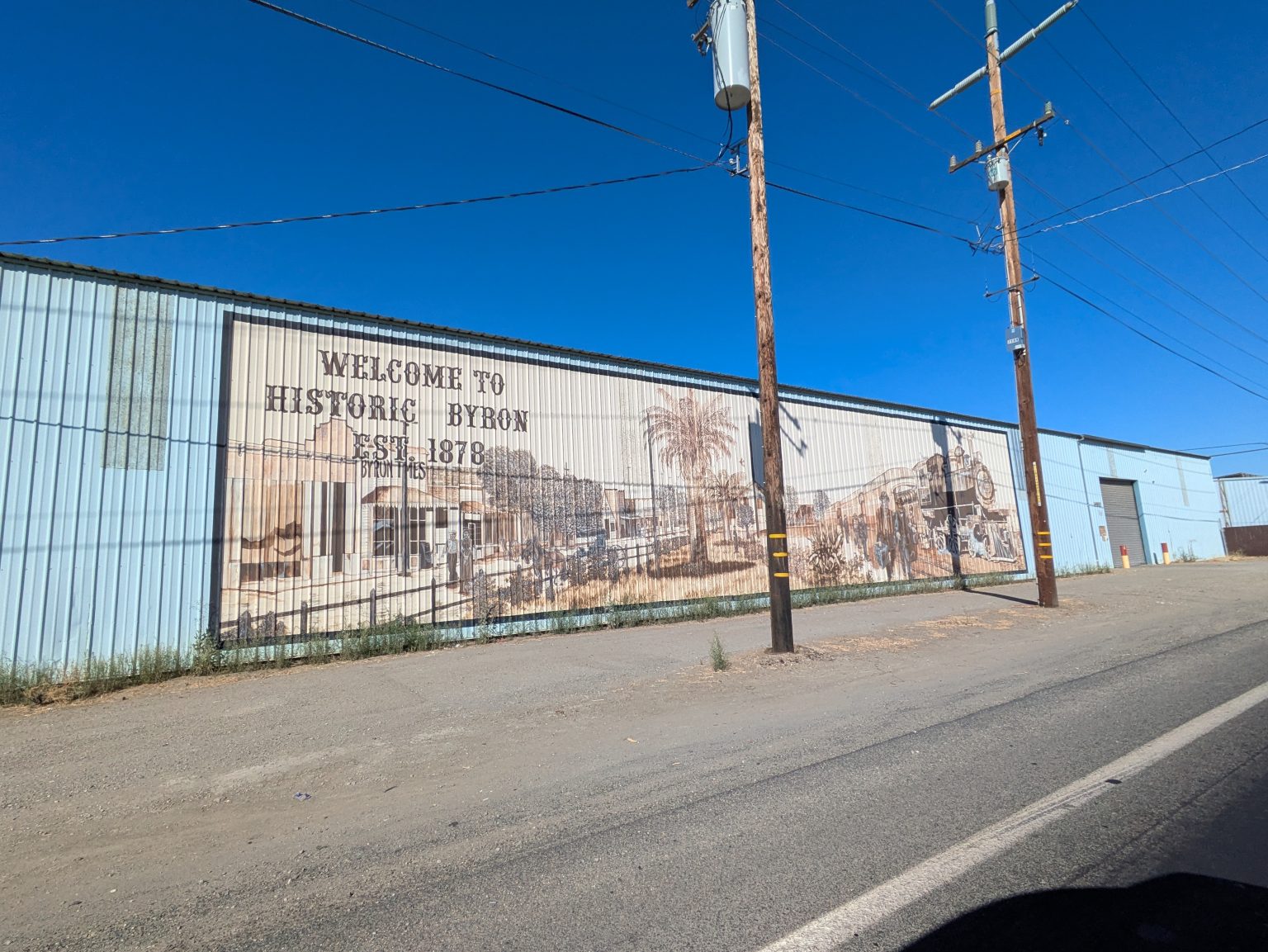 Gallery 3 - A wide outdoor mural painted in sepia tones across corrugated metal siding shows historical buildings, a palm tree, and a vintage steam train with arriving passengers, alongside text reading 
