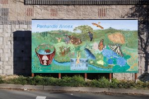 A freestanding outdoor mural mounted on wooden panels showing a colorful landscape of rolling green hills, local birds, native California plants, and historical area wildlife.