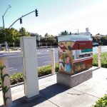 Gallery 1 - painted utility box and adjacent bollards on a sidewalk near a roadway intersection. The mural features scenes of rural life with figures, farm buildings, sacks of produce, and children seated inside an open-air classroom before a chalkboard.