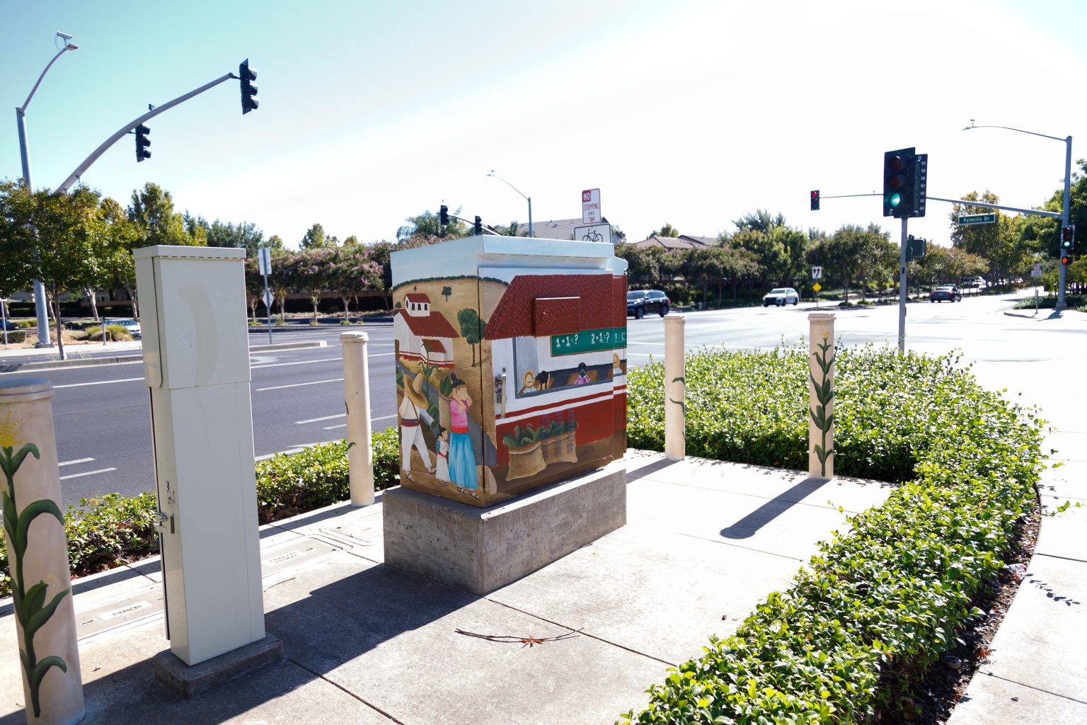 Gallery 1 - painted utility box and adjacent bollards on a sidewalk near a roadway intersection. The mural features scenes of rural life with figures, farm buildings, sacks of produce, and children seated inside an open-air classroom before a chalkboard.