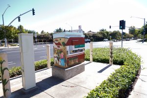 painted utility box and adjacent bollards on a sidewalk near a roadway intersection. The mural features scenes of rural life with figures, farm buildings, sacks of produce, and children seated inside an open-air classroom before a chalkboard.