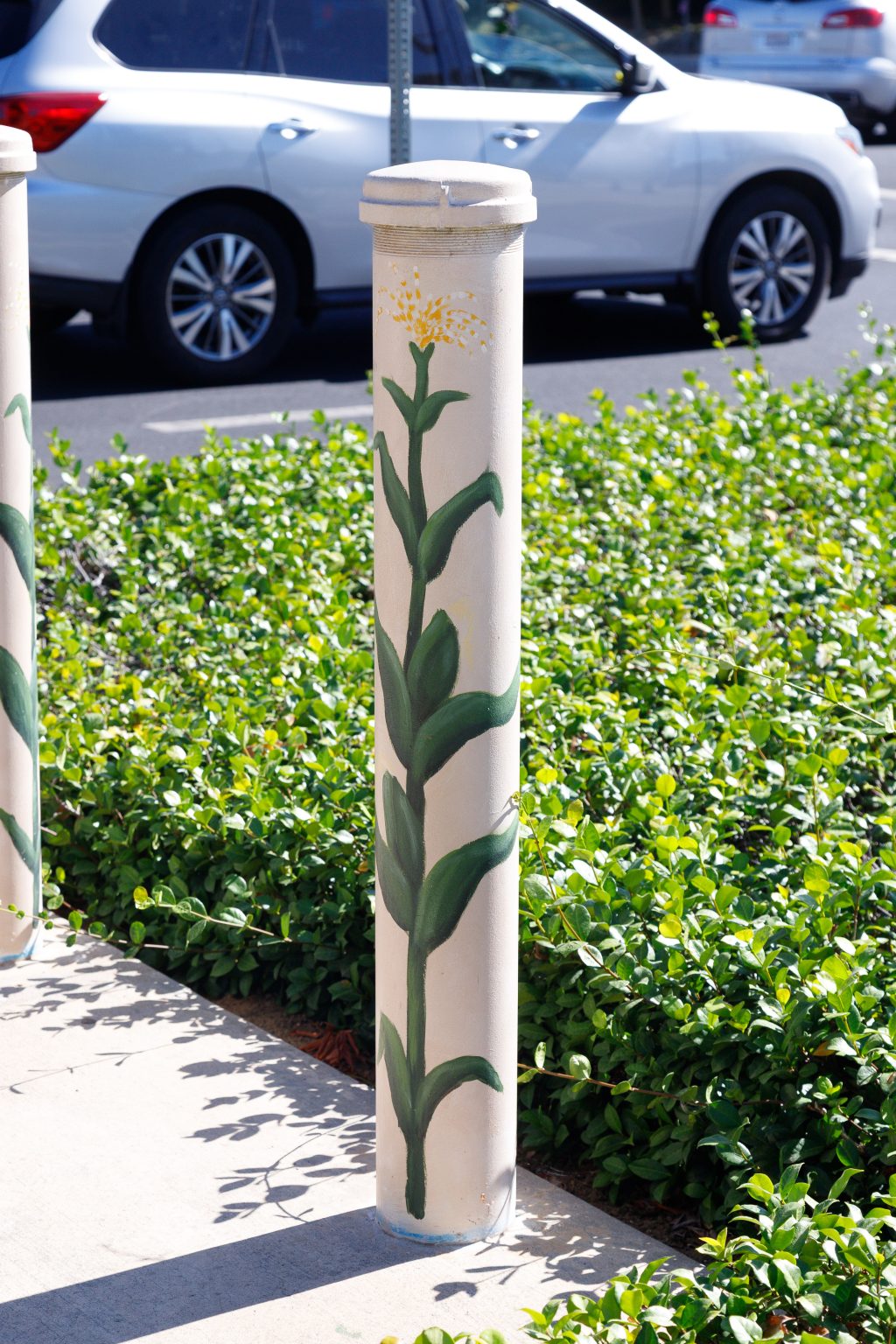 Gallery 4 - painted utility box and adjacent bollards on a sidewalk near a roadway intersection. The mural features scenes of rural life with figures, farm buildings, sacks of produce, and children seated inside an open-air classroom before a chalkboard.