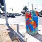 Gallery 1 - A vibrant painted utility box featuring a pink hummingbird in flight against a bright blue sky, surrounded by orange fruit and small white blossoms, with text reading 