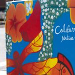 Gallery 4 - A vibrant painted utility box featuring a pink hummingbird in flight against a bright blue sky, surrounded by orange fruit and small white blossoms, with text reading 