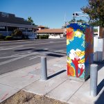 Gallery 5 - A vibrant painted utility box featuring a pink hummingbird in flight against a bright blue sky, surrounded by orange fruit and small white blossoms, with text reading 