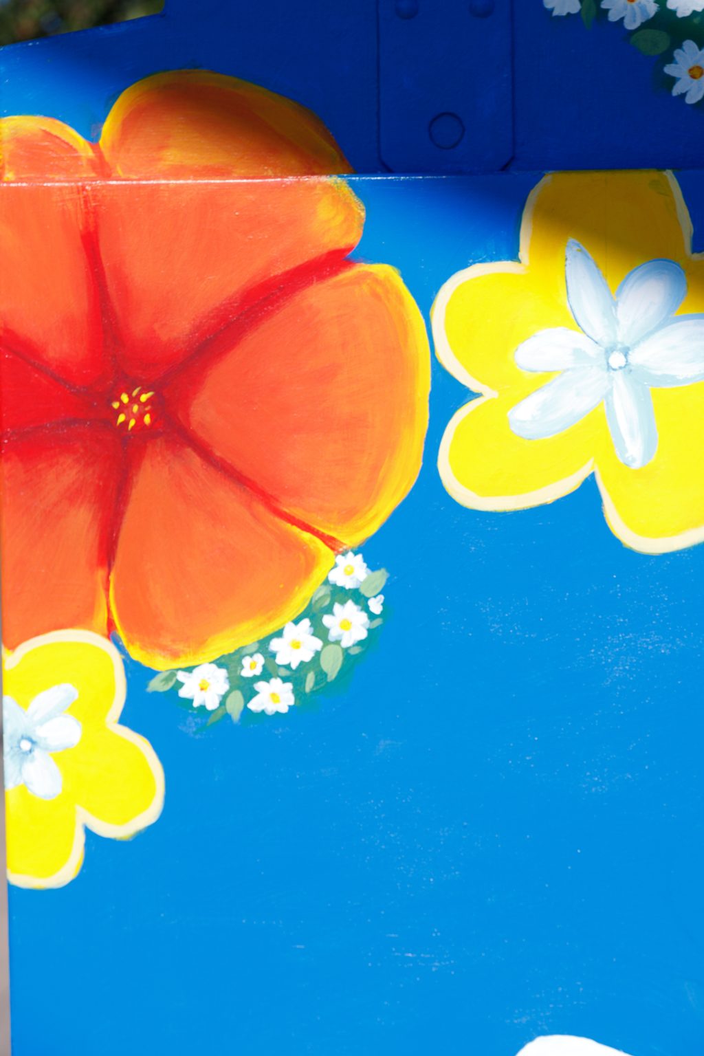 Gallery 3 - A vibrant painted utility box featuring a pink hummingbird in flight against a bright blue sky, surrounded by orange fruit and small white blossoms, with text reading 