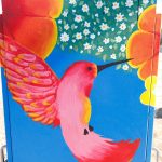 Gallery 6 - A vibrant painted utility box featuring a pink hummingbird in flight against a bright blue sky, surrounded by orange fruit and small white blossoms, with text reading 