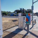 Gallery 7 - A vibrant painted utility box featuring a pink hummingbird in flight against a bright blue sky, surrounded by orange fruit and small white blossoms, with text reading 